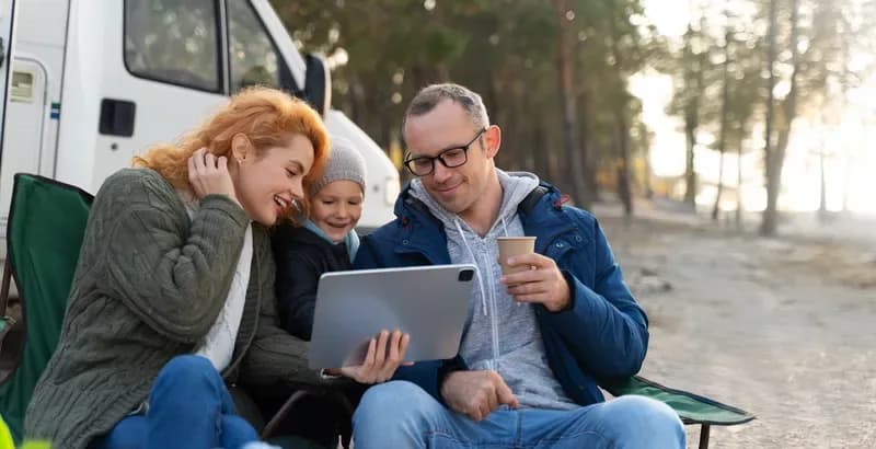 Familie sitzt in Campingstühlen vor einem Wohnmobil und schaut zusammen auf ein Tablet