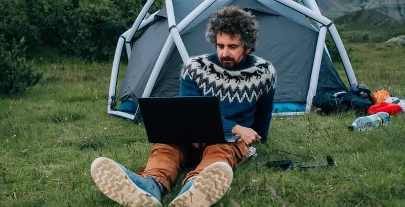 Mann sitzt mit Laptop vor einem Kuppelzelt auf einer Wiese