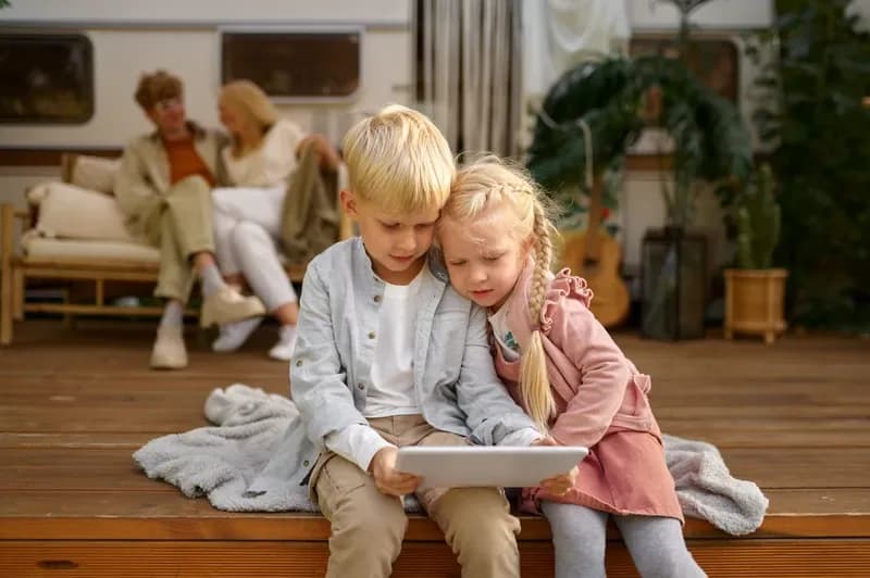 Zwei Kinder sitzen auf einer Holzterrasse vor einem Wohnwagen und schauen auf ein Tablet, im Hintergrund die Eltern