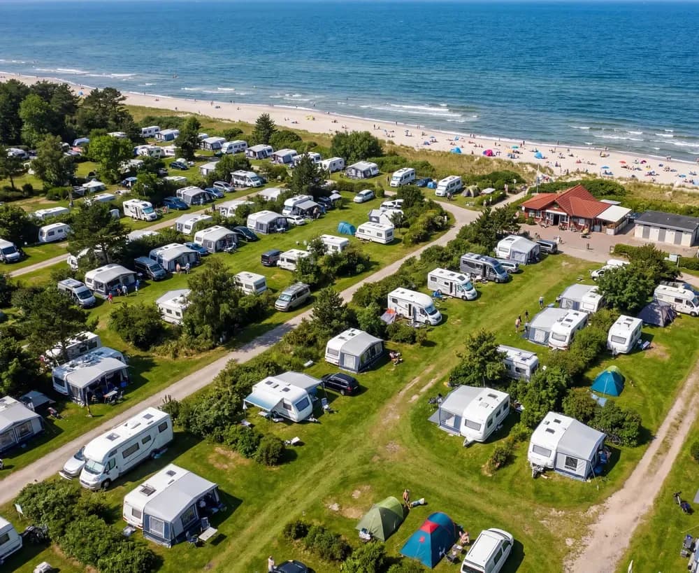 Luftaufnahme eines Campingplatzes an der Ostsee: Wohnwagen und Zelte in Reihen, dahinter Strand und Meer