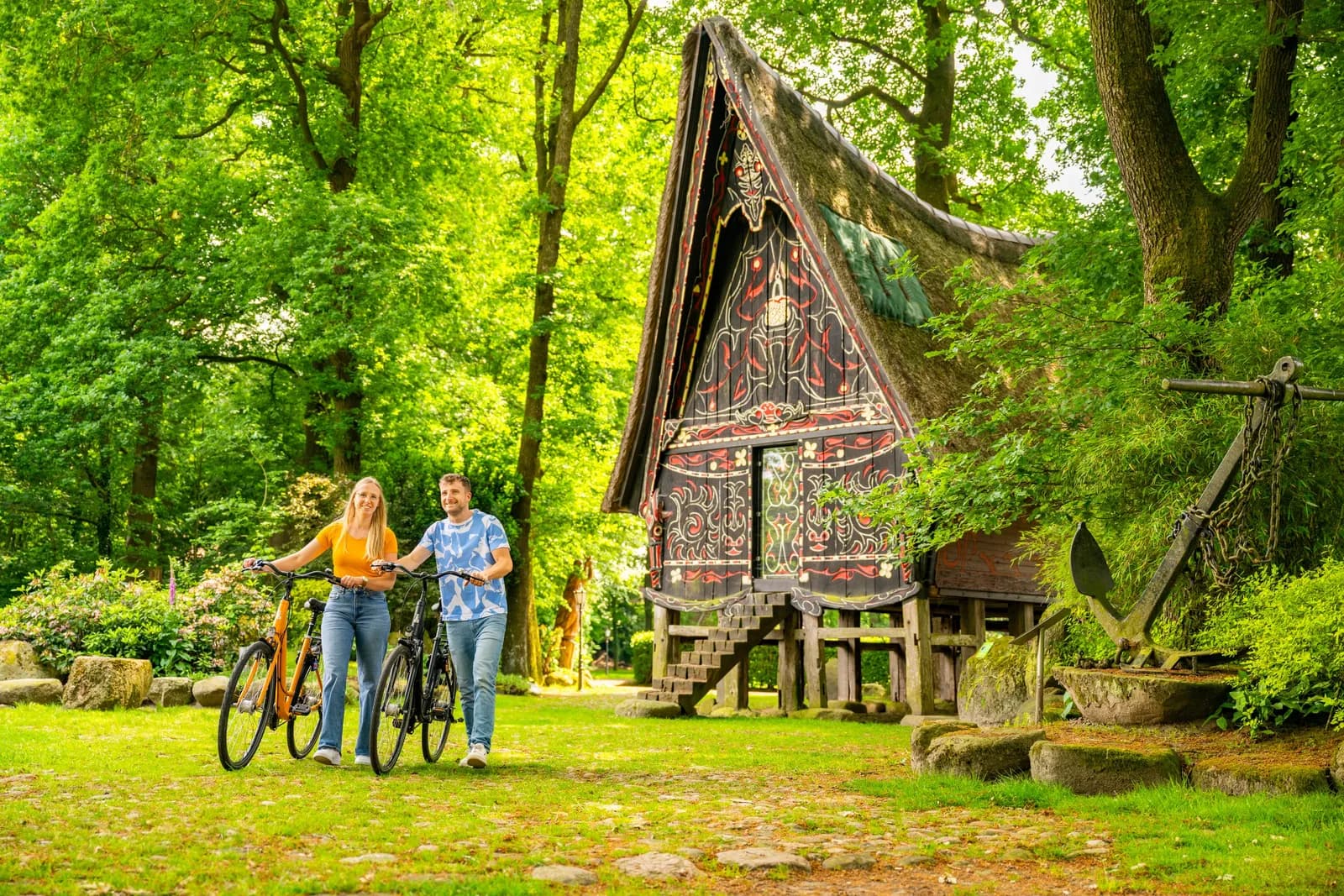 Zwei Radfahrende halten vor einem Holzhaus am Rand eines Laubwaldes