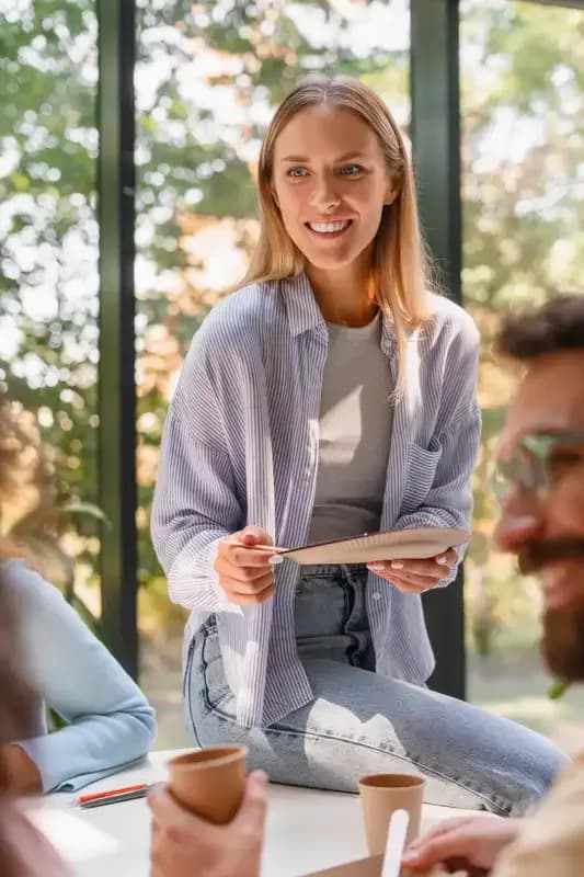 Junge Frau mit Notizblock im Gespräch mit mehreren Personen an einem Tisch vor einer Fensterfront