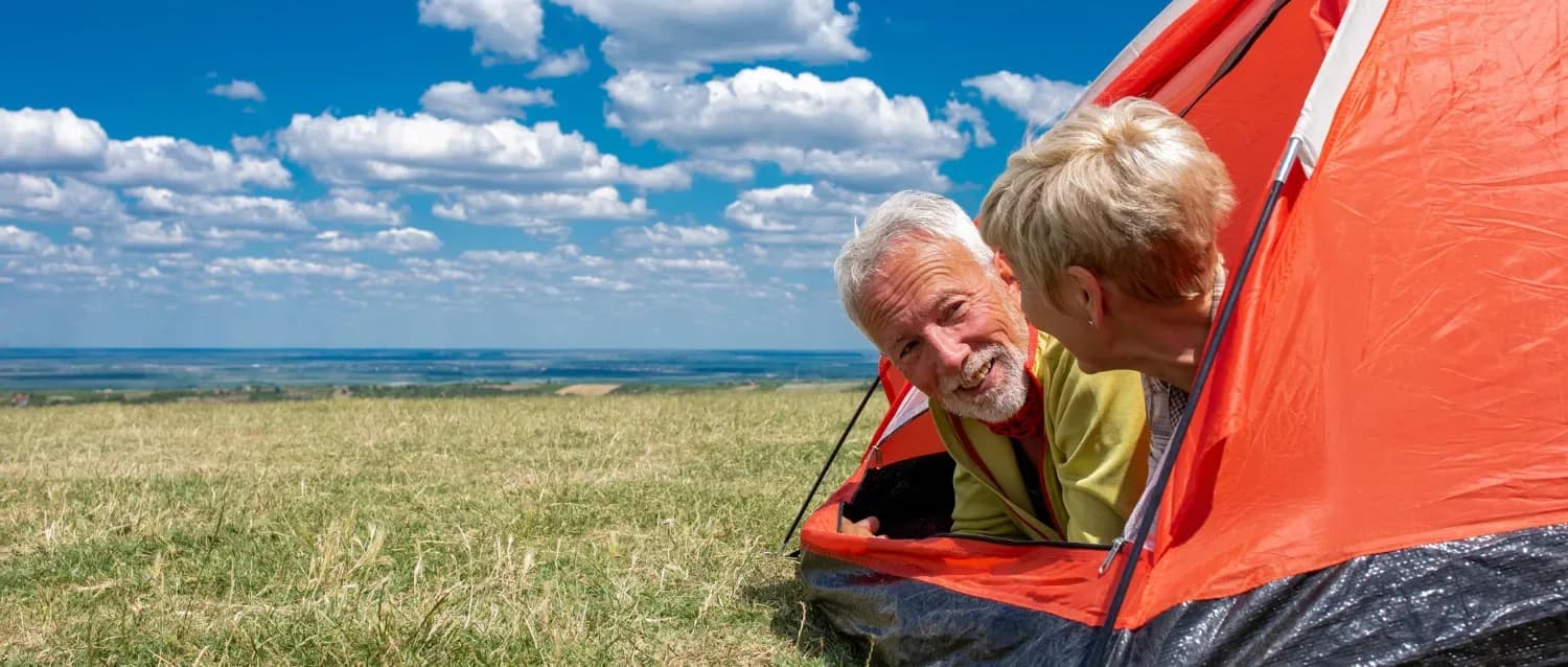 Älteres Paar schaut aus einem roten Zelt über eine weite Wiese unter blauem Himmel